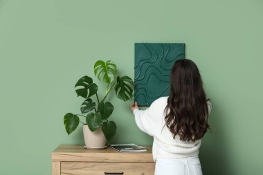 Evdeki ahşap çekmecelerin yanındaki yeşil duvara resim asan genç ve güzel bir kadın.