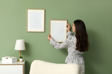 Evdeki yeşil duvara boş bir çerçeve asan genç kadın.