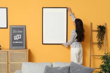 Evdeki turuncu duvara boş bir çerçeve asan genç bir kadın. Arka plan