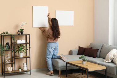 Oturma odasındaki bej duvara boş tuval asan genç bir kadın. Arka plan