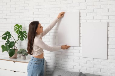 Oturma odasındaki beyaz tuğla duvara boş tuval asan genç bir kadın.