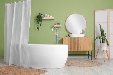 Banyonun içindeki yeşil duvarın yanındaki çekmeceleri ve perdeleri olan küvet.