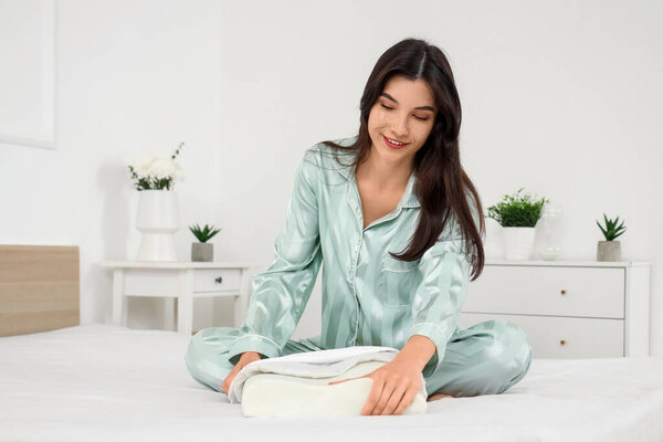 Young woman in pajamas putting cozy orthopedic pillow in pillowcase on bed at home