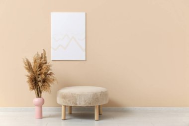 Bej duvarda asılı resim, pouf ve vazo, odada otlar var.