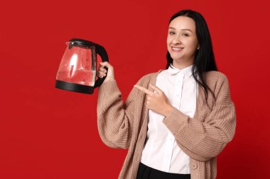 Güzel genç bayan kırmızı arka planda modern elektrikli su ısıtıcısını işaret ediyor.