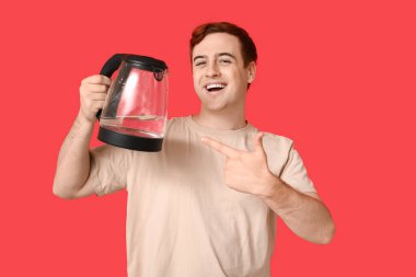 Yakışıklı genç adam kırmızı arka planda modern elektrikli su ısıtıcısını gösteriyor.