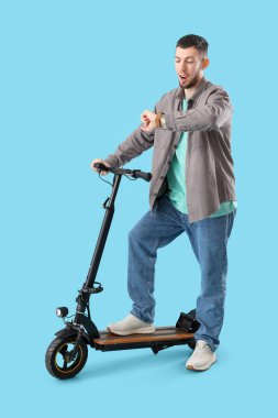 Mavi arka planda elektrikli scooterlı genç bir adam kol saatine bakıyor.
