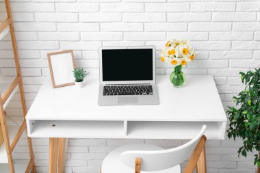 İşyerindeki beyaz tuğla duvarın yanında narsisli çiçekler ve dizüstü bilgisayarla aydınlık ofis.