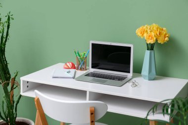 Bir buket narsisli çiçek, ev bitkileri ve dizüstü bilgisayarlı iş yeri şık bir ofiste yeşil duvarın yanında.