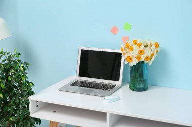 Şık bir ofiste mavi duvarın yanında bir buket narsisli çiçek ve dizüstü bilgisayarlı bir iş yeri.