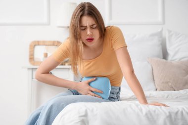 Young woman with hot water bottle suffering from menstrual cramps in bedroom