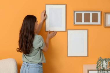 Oturma odasındaki turuncu duvara boş bir çerçeve asan genç bir kadın. Arka plan