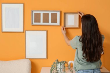 Oturma odasındaki turuncu duvara boş bir çerçeve asan genç bir kadın. Arka plan