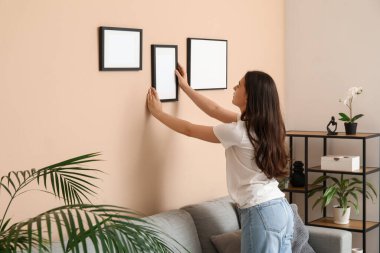 Evdeki bej duvarda boş bir çerçeve asılı genç bir kadın.