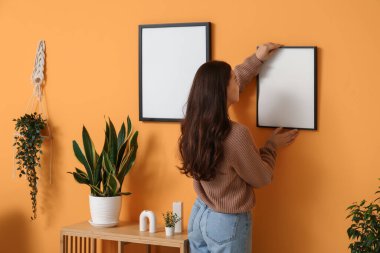 Oturma odasındaki turuncu duvara boş bir çerçeve asan genç bir kadın. Arka plan