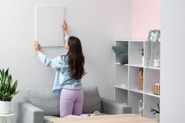Oturma odasındaki beyaz duvara boş tuval asan genç bir kadın.