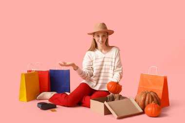 Pembe arka planda balkabağı ve alışveriş torbalarıyla sonbahar kıyafetleri içinde genç bir kadın.
