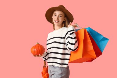 Sonbahar kıyafetleri içinde, balkabağı, şemsiye ve pembe arka planda alışveriş torbalarıyla genç bir kadın.