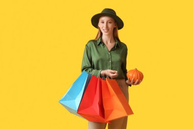 Sarı arka planda balkabağı ve alışveriş torbalarıyla sonbahar kıyafetleri içinde genç bir kadın.