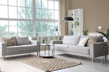 Oturma odasının içi rahat gri koltuklar, kahve masası ve büyük pencereli.