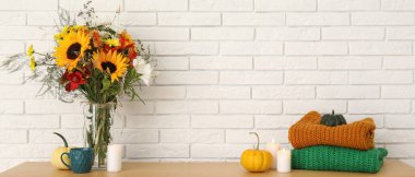 Sonbahar çiçekleri, süveterler, balkabakları ve beyaz tuğla duvarın yanında yanan mumlarla dolu ahşap masa.