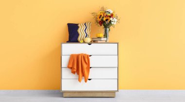 Sonbahar çiçekleri ve sarı duvarın yanında balkabakları olan beyaz çekmeceler.