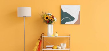 Sonbahar çiçekleri, standart lamba ve şemsiyeli raflar sarı duvarın yanında.