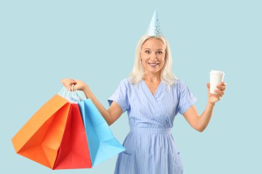 Parti şapkalı olgun bir kadın. Elinde kahve fincanı ve mavi arka planda alışveriş torbaları var.