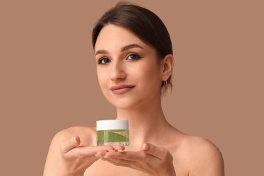 Kahverengi arka planda kozmetik kremi kavanozu olan güzel genç bir kadın.