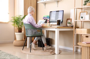 Gözlüklü genç bayan moda tasarımcısı hafif ofisteki bilgisayarla masada oturuyor.