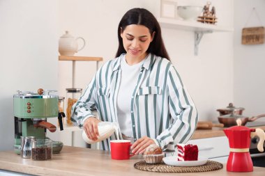 Güzel, genç, mutlu bir kadın mutfakta bir fincan taze kahveye süt dolduruyor.