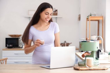 Mutfakta bir fincan cappuccino ve dizüstü bilgisayarlı güzel, mutlu bir kadın.