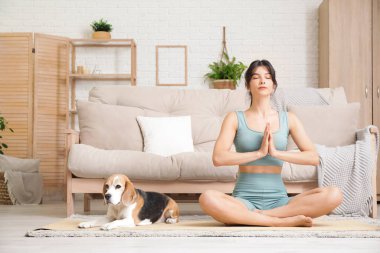 Genç sporcu kadın ve Beagle köpeği oturma odasında yoga minderi üzerinde meditasyon yapıyorlar.