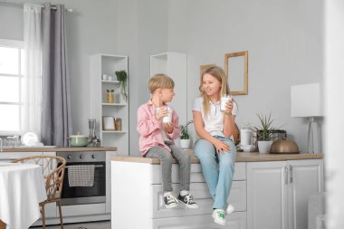 Mutfaktaki tezgahta süt bardaklarıyla oturan iki kardeş.