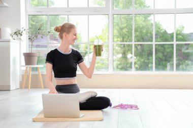 Sağlıklı smoothie ve dizüstü bilgisayarlı genç bir kadın mutfakta yoga minderinde oturuyor.