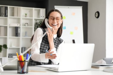 İş yerinde modern dizüstü bilgisayarla şık bir ofiste telefonla konuşan genç bir kadın.