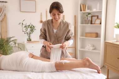 Masaj terapisti kadın bacağını kaplıca salonundaki kanepeye bandajlıyor.