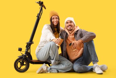 Kahve fincanları ve elektrikli scooterlı genç çift sarı arka planda oturuyor.