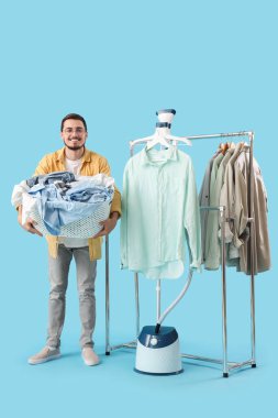 Modern buharlı ve çamaşır dolu sepetli mutlu genç adam mavi arka plandaki elbise rafının yanında.