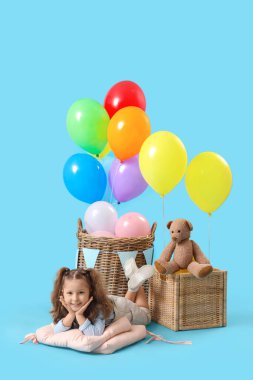Sevimli küçük kız mavi arka planda balonları ve oyuncakları olan sepetlerin yanında.
