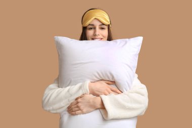 Pijamalı ve uyku maskeli genç bir kadın bej arka planda yastık tutuyor.