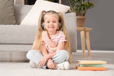 Oturma odasında halıda oturmuş kitaplarla sesli kitap dinleyen kulaklıklı tatlı küçük kız.