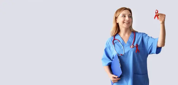 stock image Mature female doctor with red ribbon on grey background. AIDS awareness concept