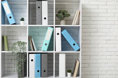 Beyaz tuğla duvarın yanında doküman dizinleri ve bitkileri olan raf ünitesi