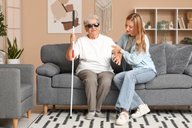 Genç bir kadın kör büyükannesinin evdeki kanepeden kalkmasına yardım ediyor.