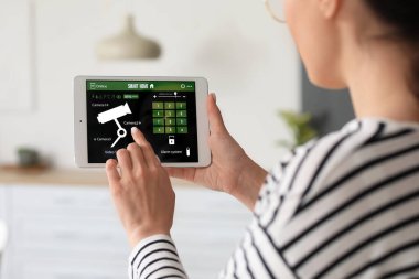 Mutfakta akıllı ev güvenlik sistemi kontrol paneli kullanan genç bir kadın.