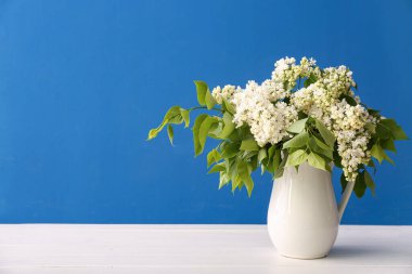 Mavi duvarın yanındaki ahşap masada beyaz leylak vazosu.