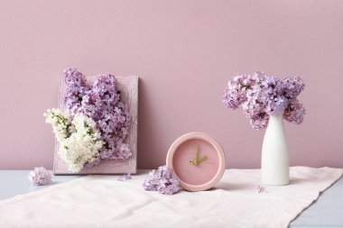 Pembe duvarın yanındaki beyaz masada çerçeveli ve saatli güzel kokulu leylak çiçekleri vazosu.