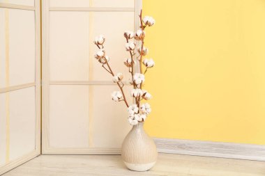 Sarı duvarın yanında giyinme ekranı olan yerde pamuk filizi vazosu var.