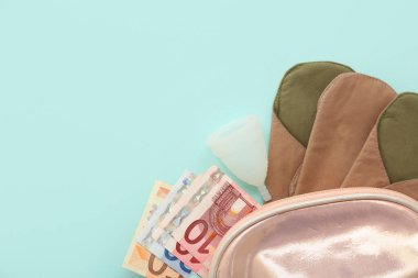 Yeniden kullanılabilir regl pedi, bardak ve turkuaz arka planda parası olan kadının makyaj çantası.